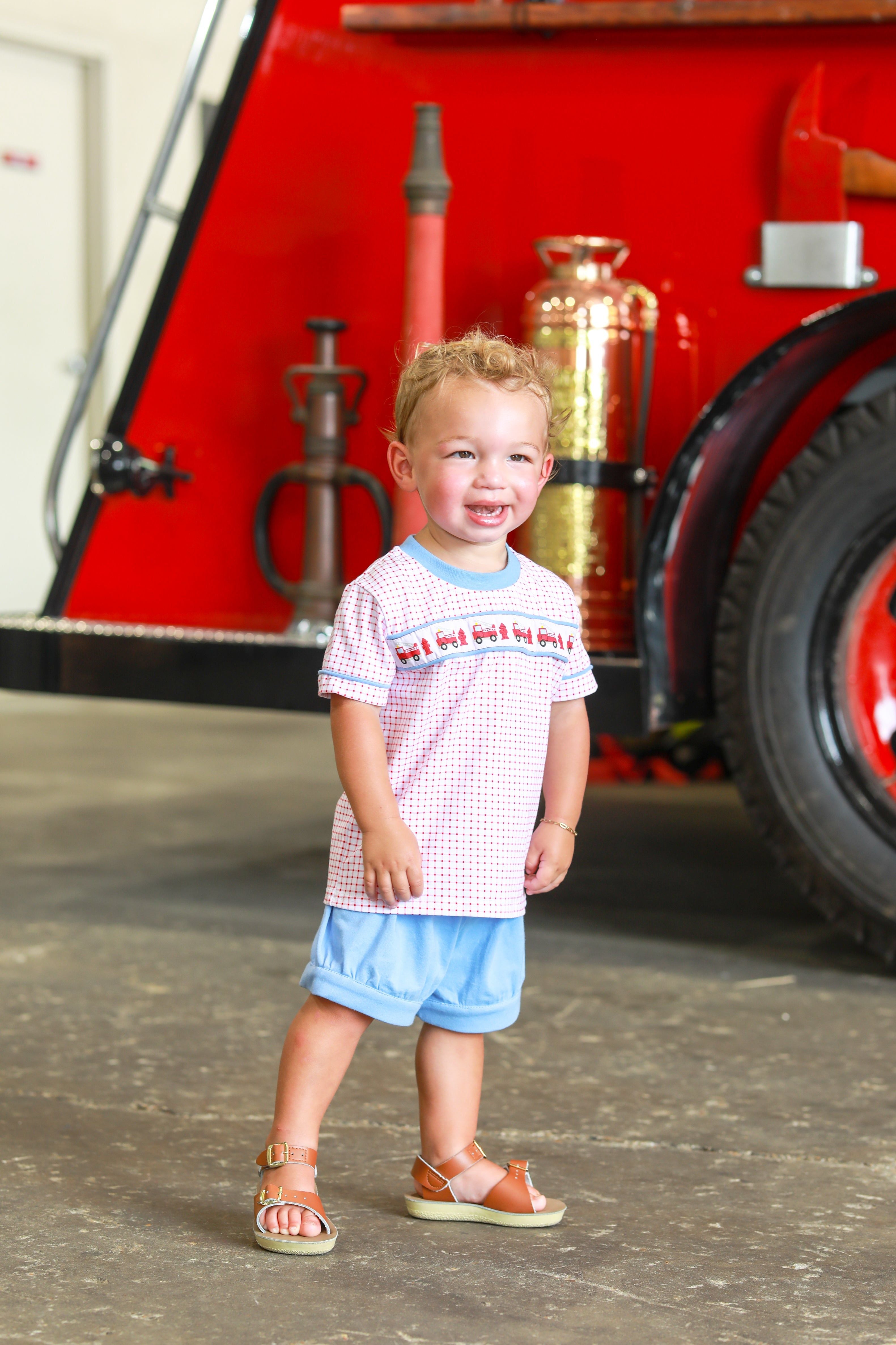 Toddler wearing red fire truck patterned shirt with blue shorts standing in front of fire truck