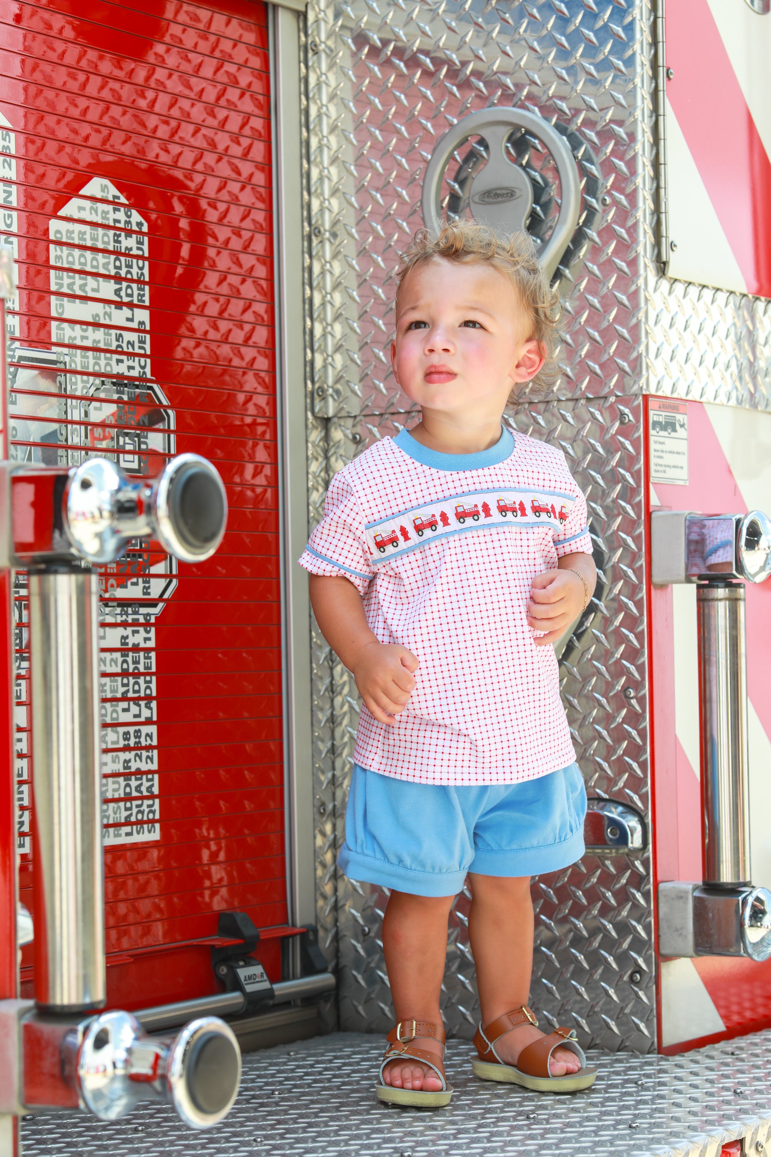 Toddler wearing red and blue fire truck patterned short set while standing on fire truck steps.