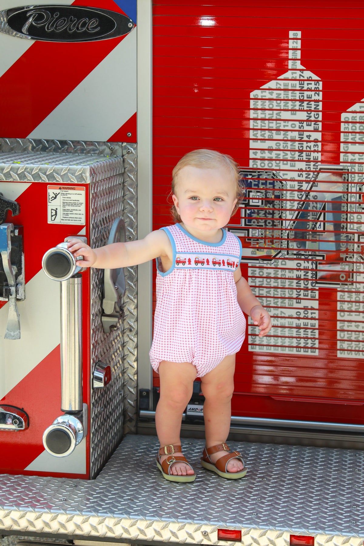 Young child in red checkered fire truck bubble romper standing on Pierce fire engine