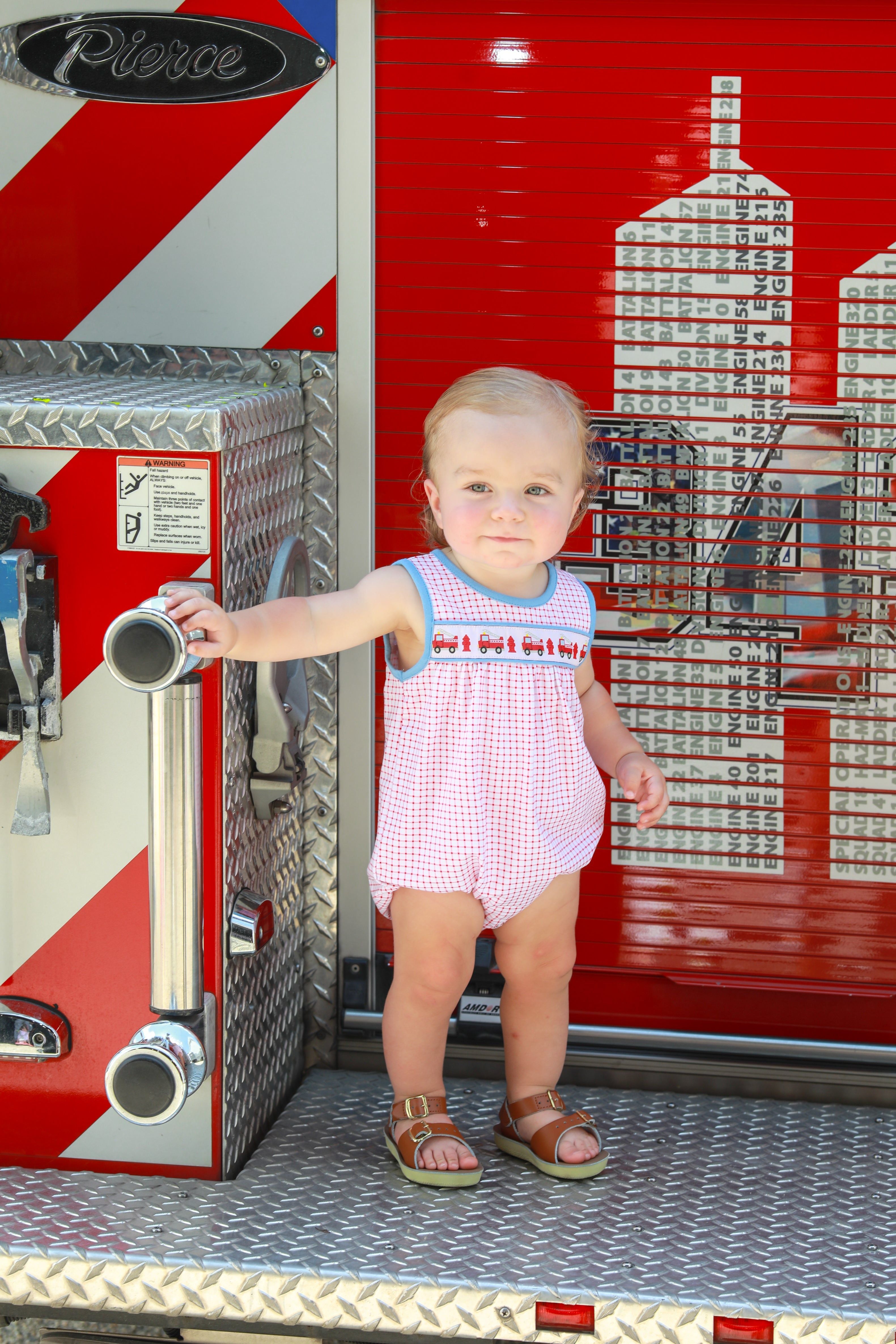 Young child in red checkered fire truck bubble romper standing on Pierce fire engine