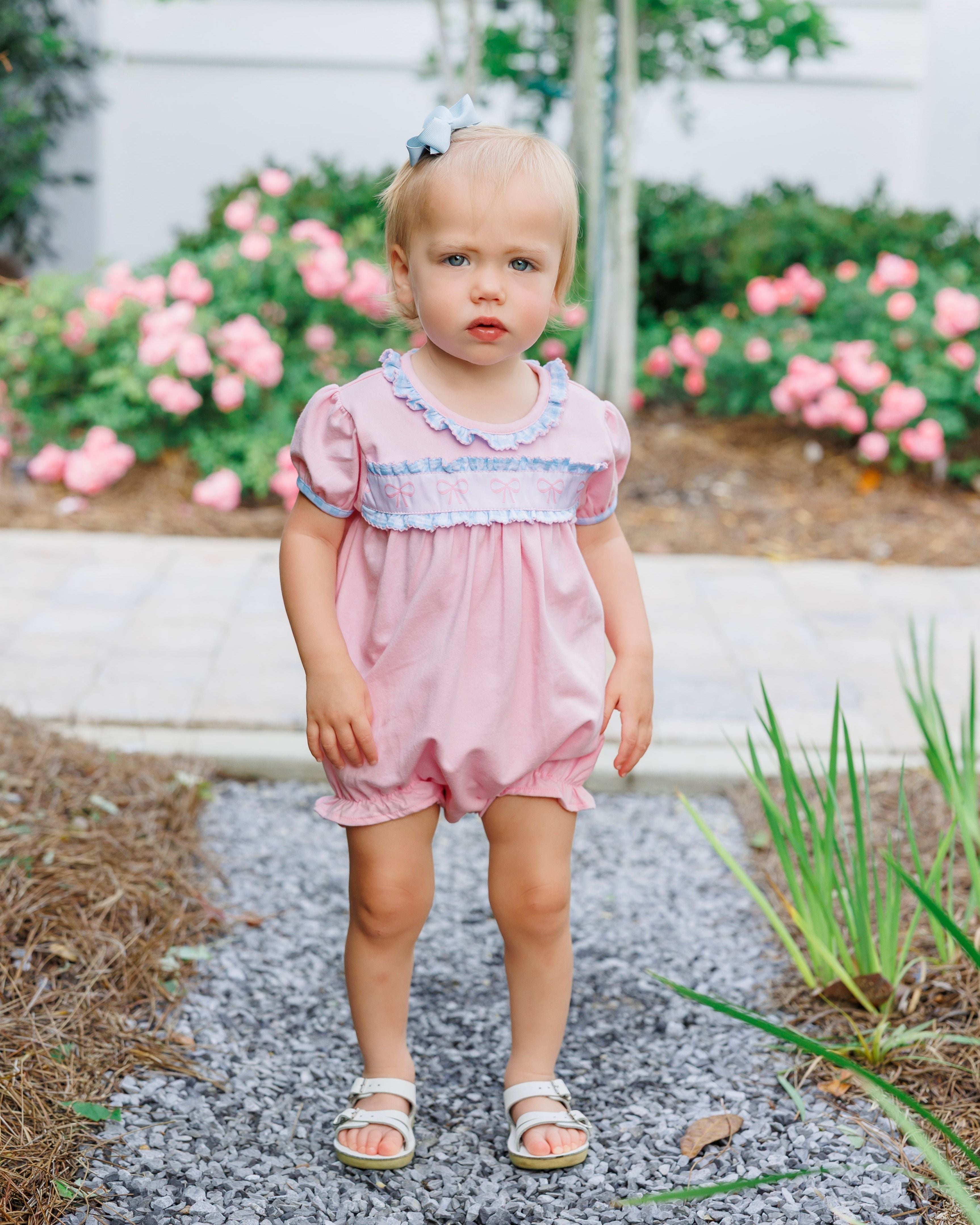Toddler wearing pink bubble romper with blue trim and matching hair bow standing on garden path