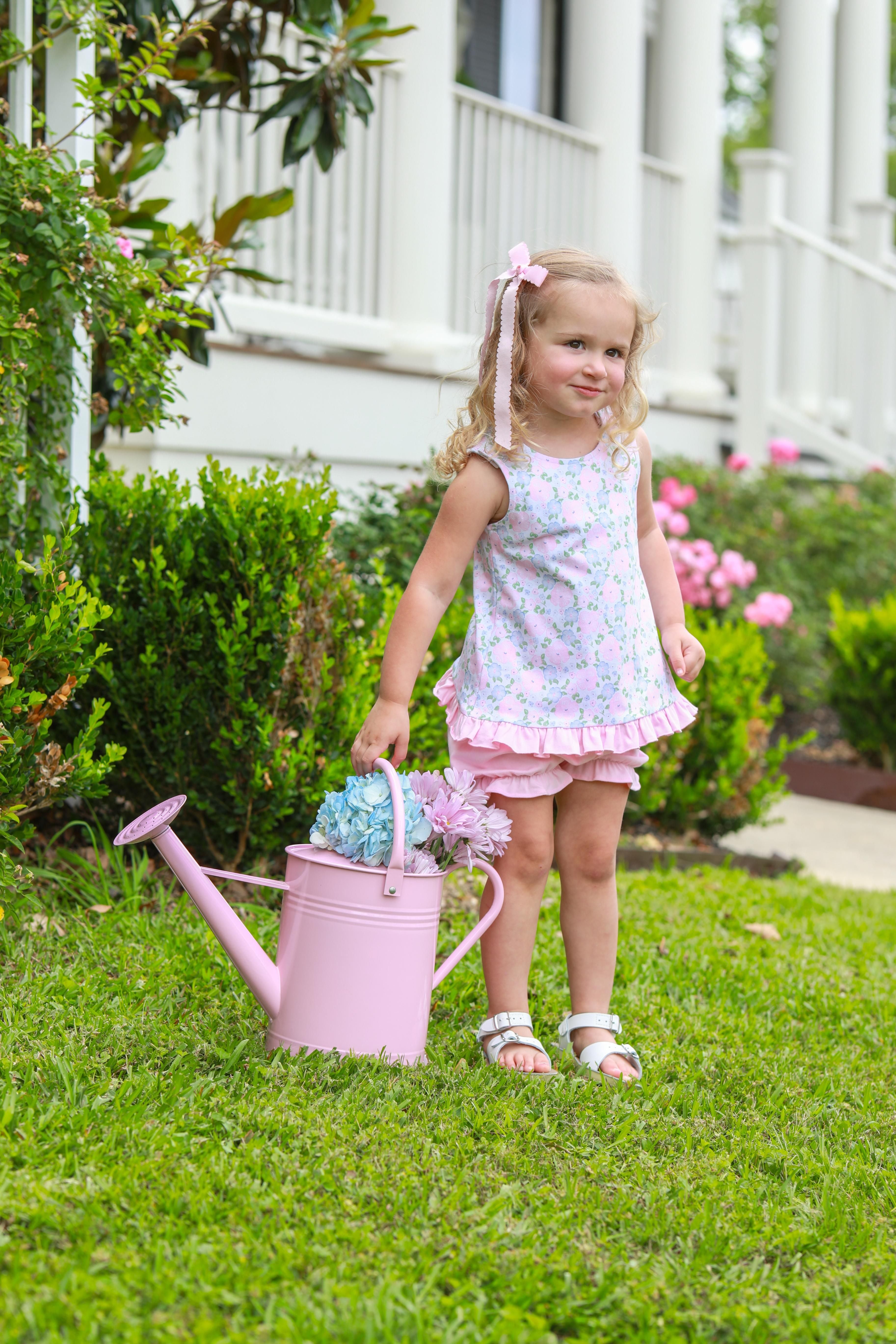 Young girl wearing pink floral sleeveless dress with ruffled bloomers, standing in garden with pink watering can.