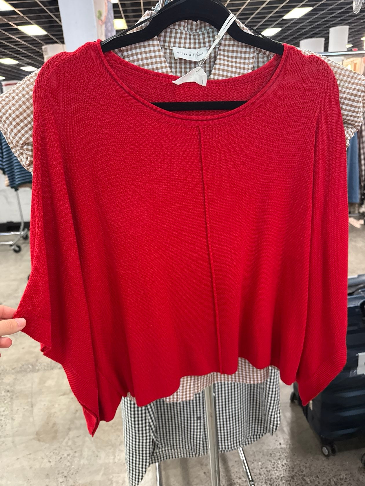 Red knit poncho-style top with draped sleeves displayed on hanger in retail store setting