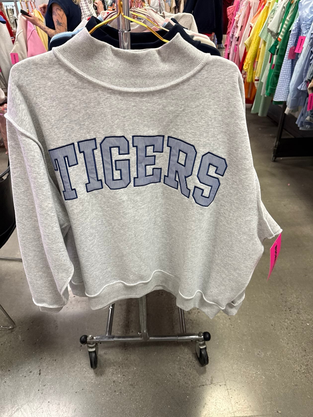 Gray cropped sweatshirt with "TIGERS" text in navy blue lettering, displayed on clothing rack in store