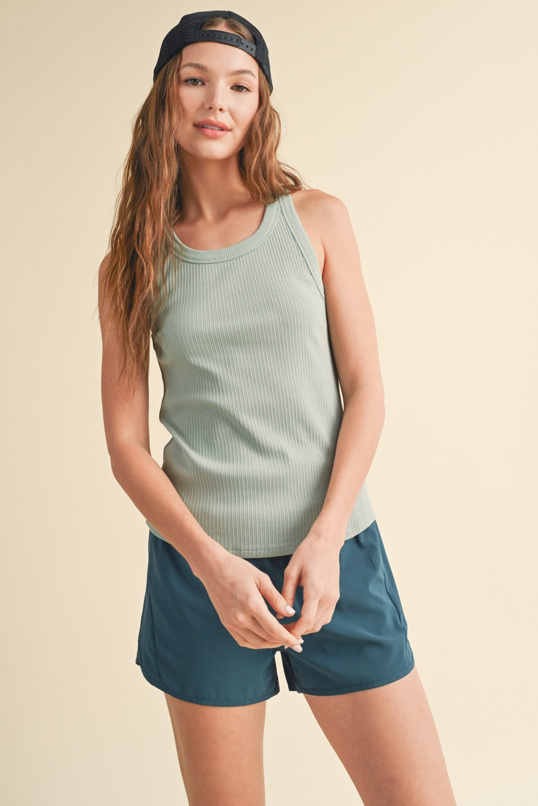 Woman wearing sage green ribbed tank top and navy blue shorts with backwards baseball cap