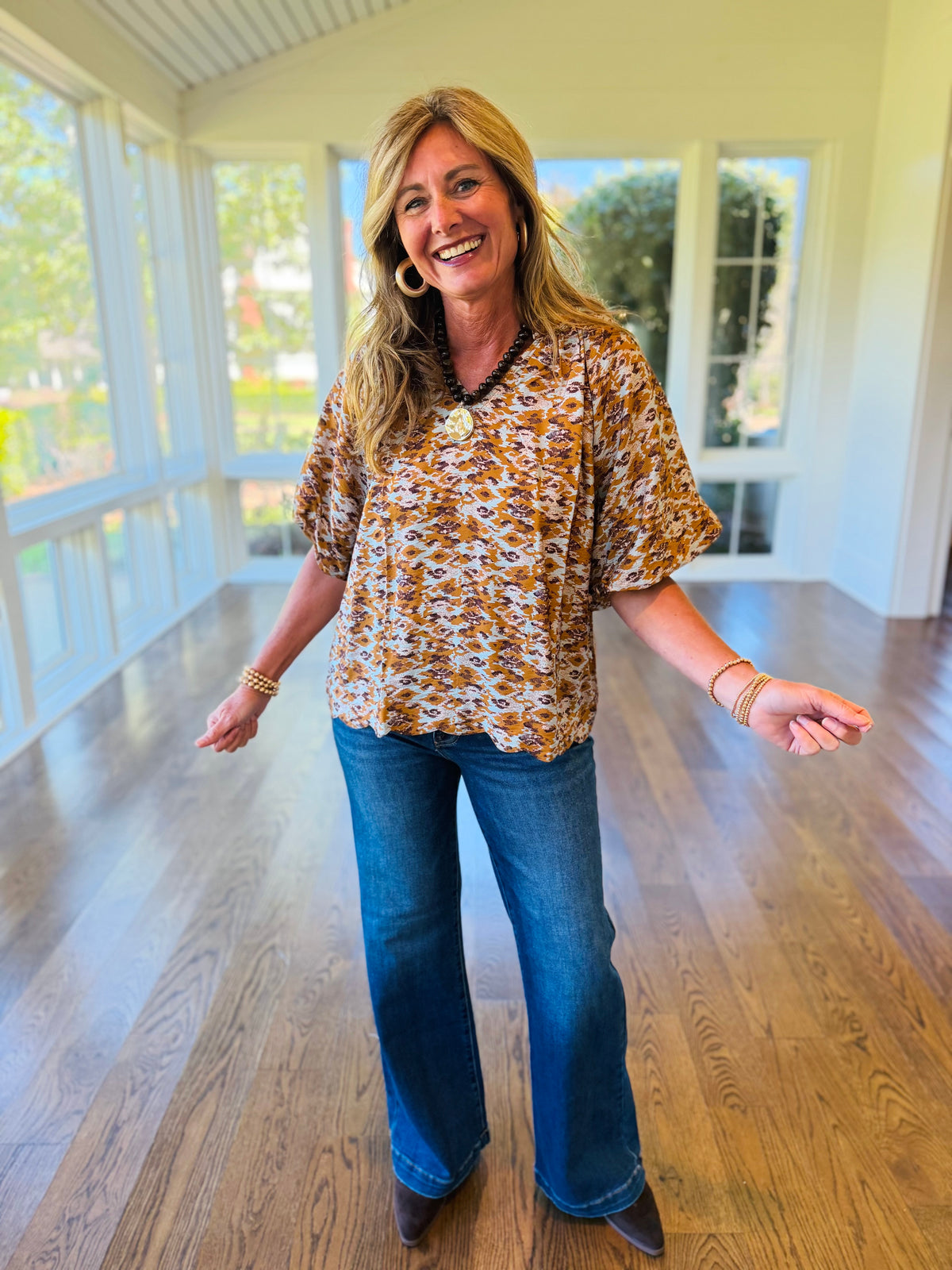 Kelly wearing the Alana rust abstract print puff sleeve blouse with blue flared jeans and dark ankle boots in a bright sunroom.