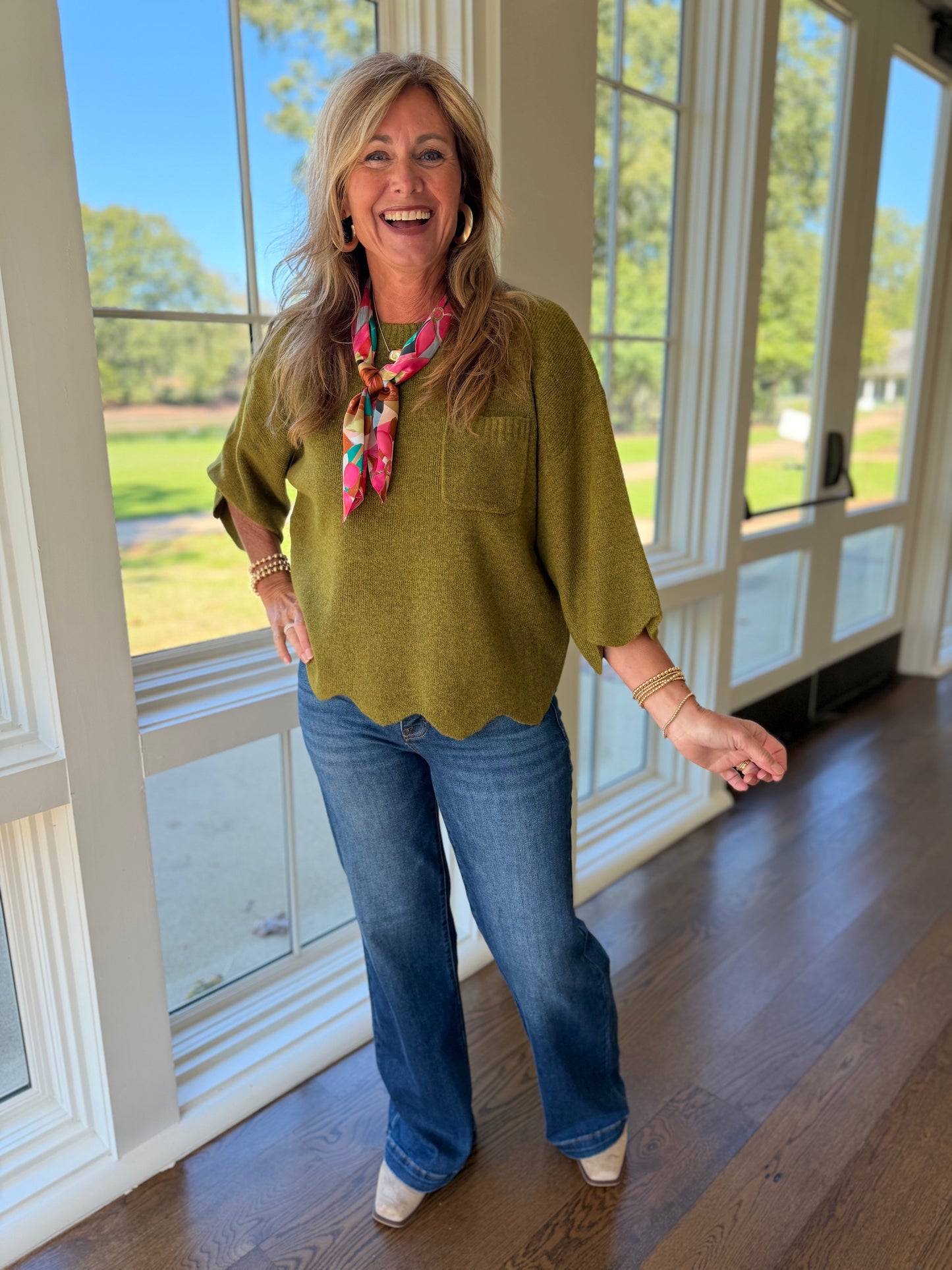 Kelly wearing the Briar olive scallop hem knit top with blue jeans and a colorful neck scarf.