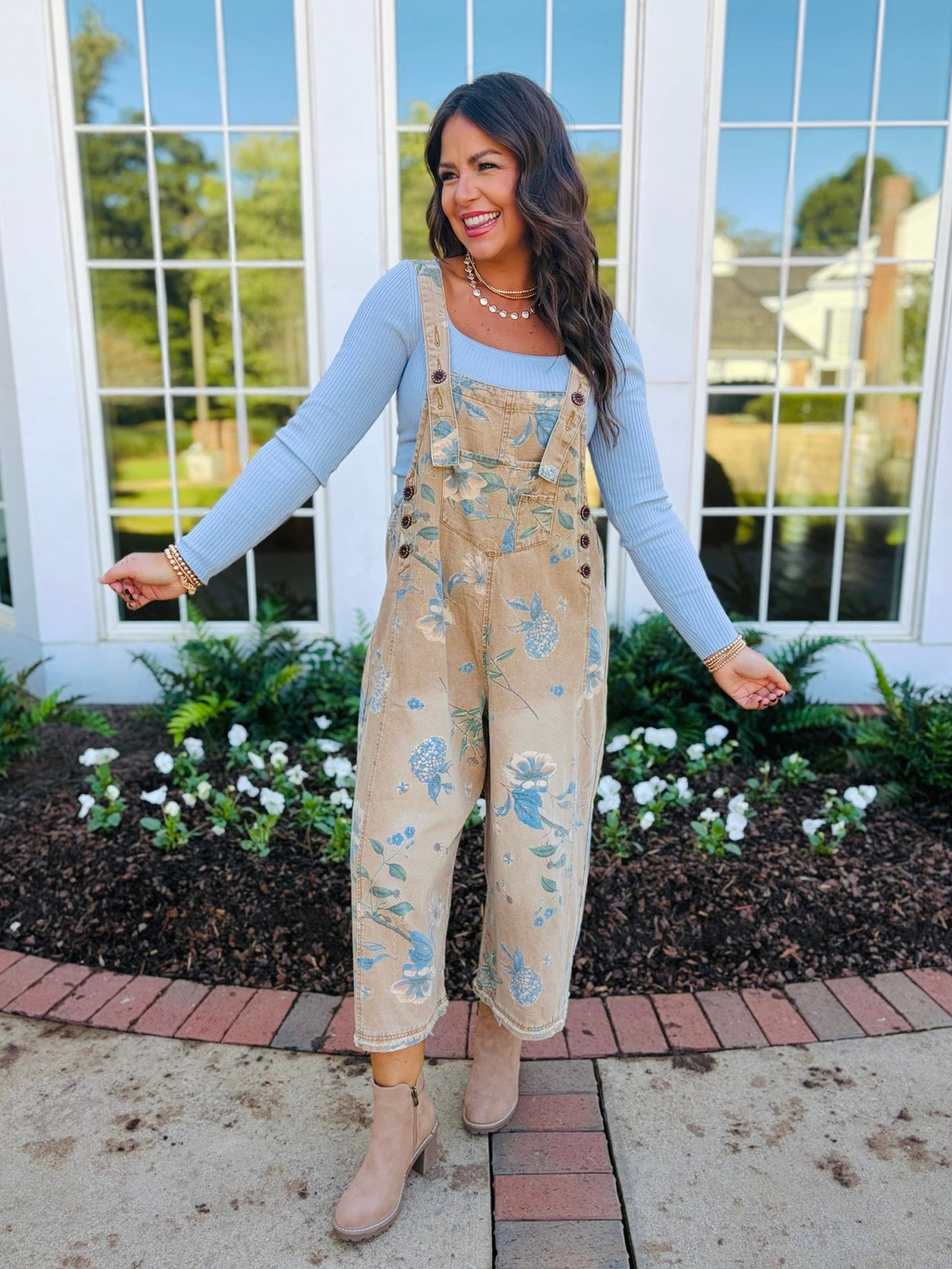 Brooke wearing the Lucille mocha floral print overalls layered over a light blue ribbed top with neutral ankle boots.
