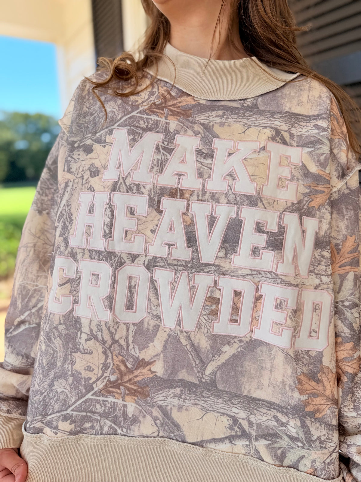Close-up of the Make Heaven Crowded camo sweatshirt showing its varsity-style lettering, mock neckline and woodland print.