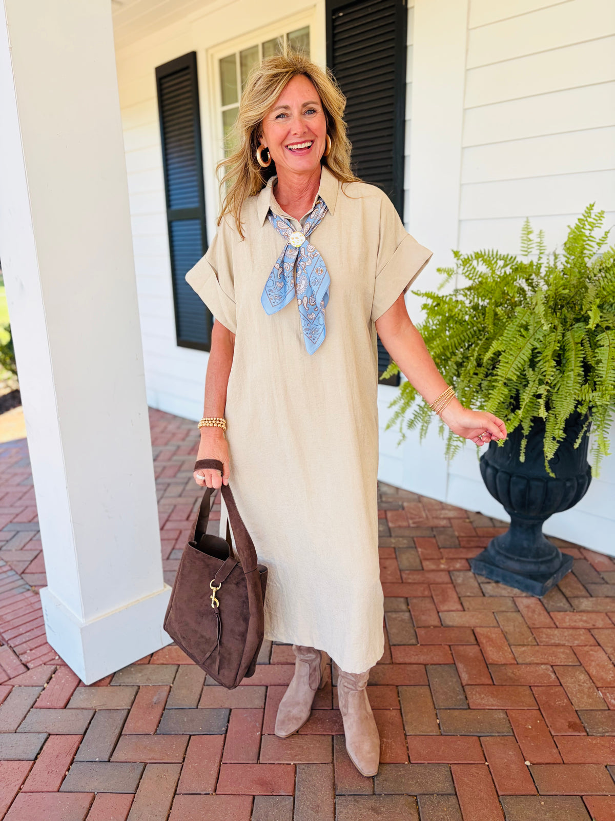 Kelly wearing the Page light taupe collared midi dress with a blue patterned scarf, brown handbag and taupe boots on a brick porch.