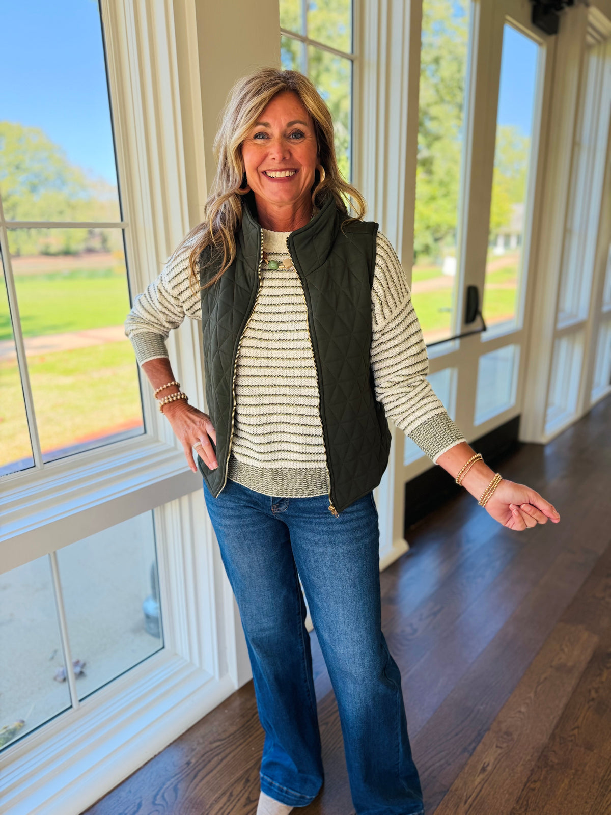 Kelly wearing the Rowan dark olive longline quilted vest over a cream striped sweater with blue jeans.