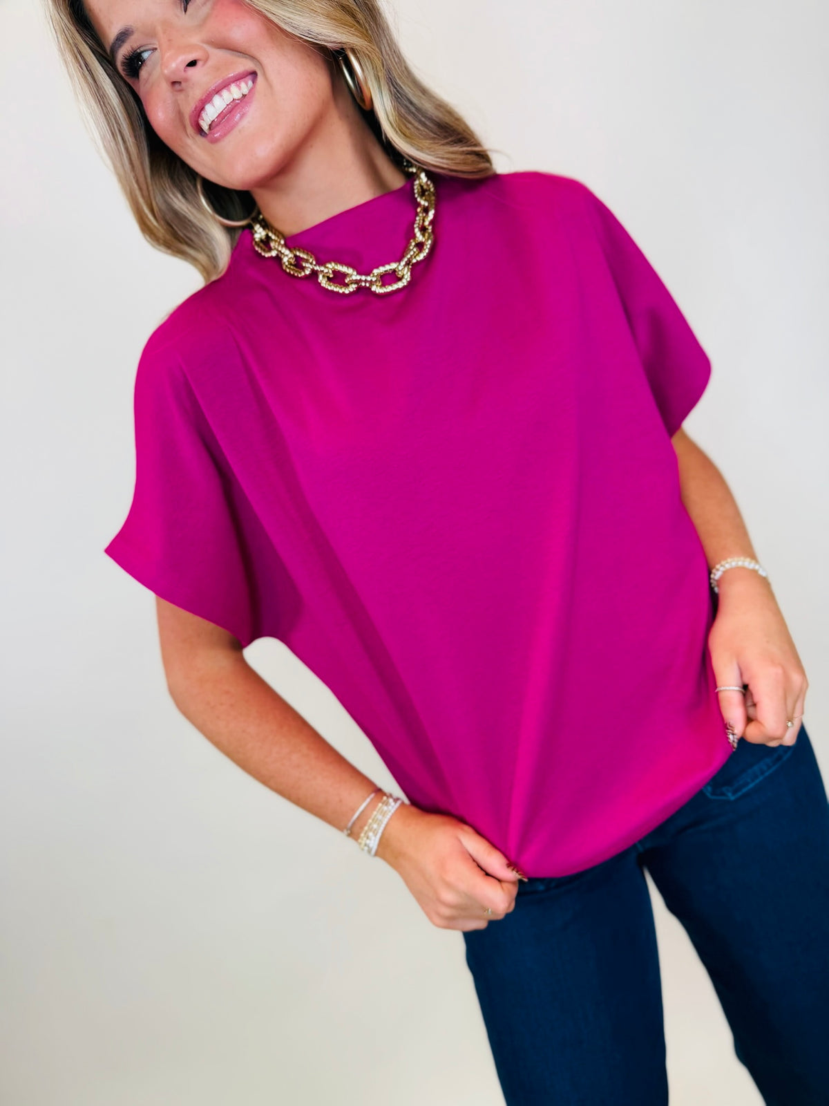 Model wearing the Sunrise magenta short sleeve high neck blouse with dark blue jeans and gold jewelry.